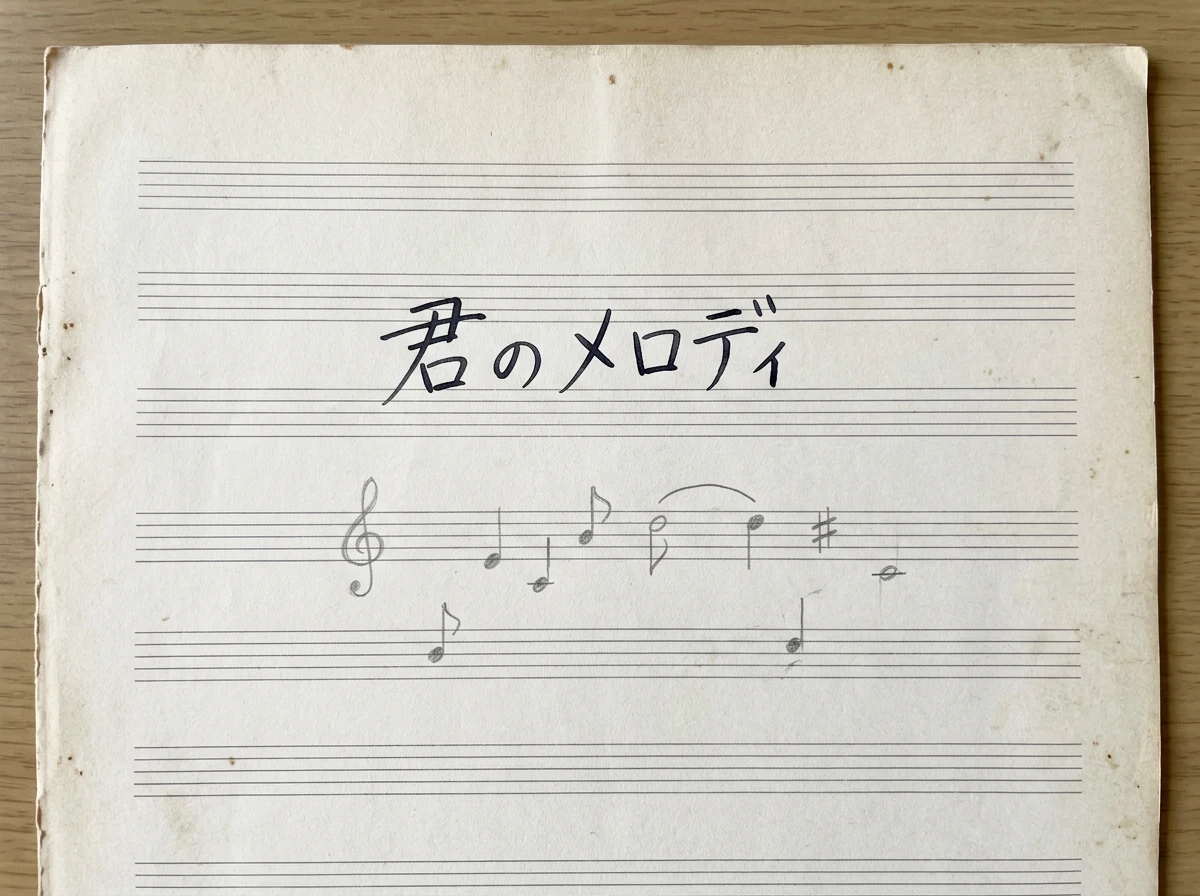 君のメロディの手書き歌詞を五線譜、音符、鉛筆メモのあるアイボリー紙に配置した音楽原稿